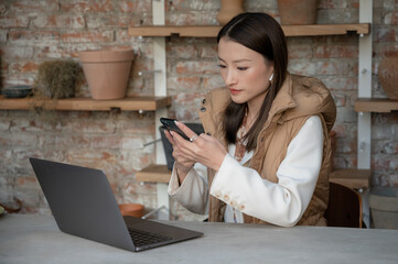 Portrait of pretty young Chinese Asian businesswoman wearing classic clothes in cafe restaurant office uses her mobile phone and laptop. Freelance worker using internet in coffee shop to make the job.
