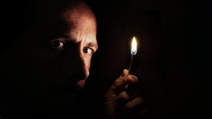 Worried man looking at the camera with a match in the dark. Blackout concept.