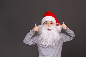 Fototapeta na wymiar a surprised Santa boy with a beard and a Santa hat on a gray background. gives a thumbs up. copy space