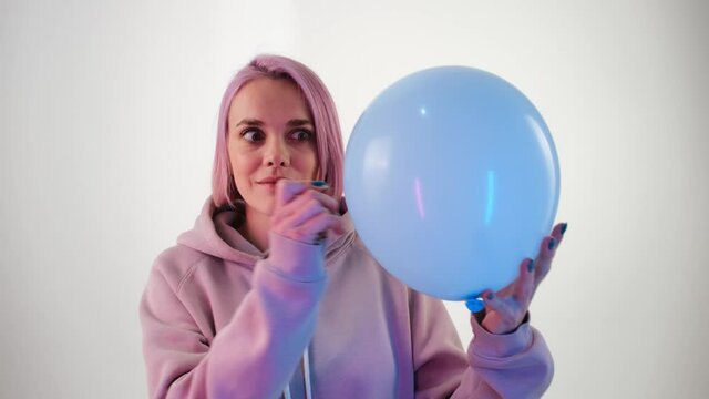 Young woman with pink hair bursting a balloon. Slow motion shoot of girl piercing with needle pink air balloon and showing surprised emotion white background