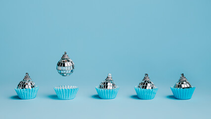 Selection of silver Christmas disco ball ornaments in a paper cupcake mold with flying one against pastel blue background. Creative New Year or Xmas celebration party concept. Festive food idea.
