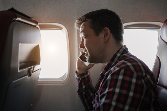 man in plaid shirt talks on phone in seat of passenger on board of plane.