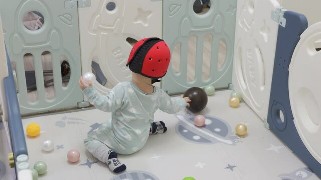 Cute baby toddler playing with ball pits in foldable baby playpen for babies and toddlers, kids activity centre safety play yard keeping child away from hurt. Baby boy wearing safety helmet.