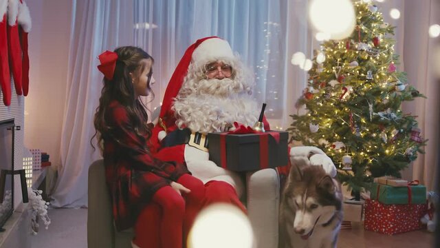 Little cute girl is sitting near fireplace on Santa's Claus knees, whispering a wish on Santa's ear. Whitebearded Santa is hugging her and holding a gift.