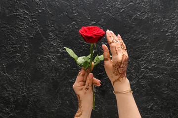 Beautiful female hands with henna tattoo holding red rose on black textured background
