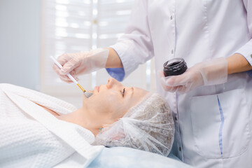 Side view of hands of cosmetology specialist applying carbon nanogel on young woman patient face using brush, getting ready for carbon peeling procedures