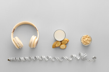 Glass of protein shake, bowl with powder, sliced banana, measuring tape and headphones on light background