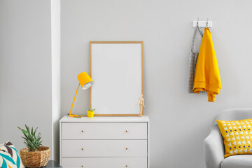 Chest of drawers with lamp, wooden mannequin and blank frame near light wall