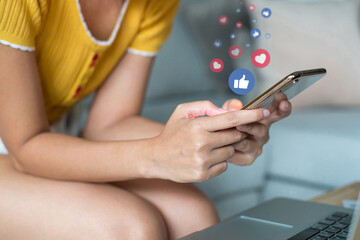 Young woman wearing yellow t-shirt sitting on sofa using smart phone to social media with Like and Love icons.