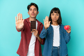 Young mixed race couple holding apple and chocolate isolated on blue background making stop gesture denying a situation that thinks wrong