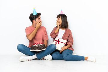 Young mixed race couple celebrating a birthday sitting on the floor isolated on white background covering mouth with hands for saying something inappropriate