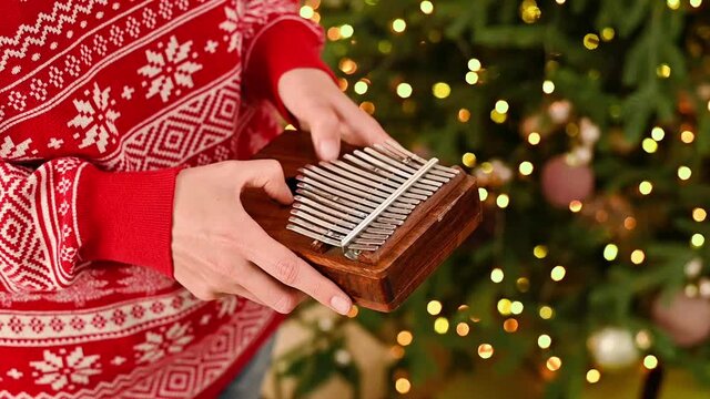 Playing on the African instrument Kalimba or Mbira on christmas tree background in red sweater. Traditional instrument from Africa close up playing christmas songs.