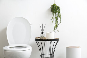 Toilet bowl, table with bathroom accessories, bin and houseplant near white wall