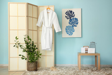 Fototapeta na wymiar Folding screen with bathrobe, table and frames near blue wall