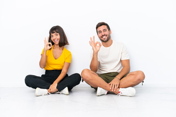 Fototapeta na wymiar Young couple sitting on the floor isolated on white background showing an ok sign with fingers