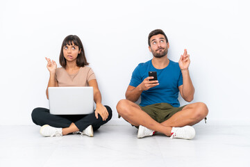 Young couple sitting on the floor holding pc and mobile phone isolated on white background with fingers crossing and wishing the best