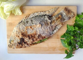 Authentic thai grilled fish (nile tilapia) on a wooden cutting board, garnished with coriander, basil and Chinese cabbage.