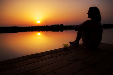 Silhouette of a woman sitting on the wooden dock, relaxing with a cup of coffee and enjoying a golden sunset over the water.