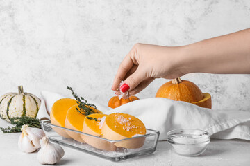 Woman adding salt in baking dish with fresh pumpkin pieces on light background
