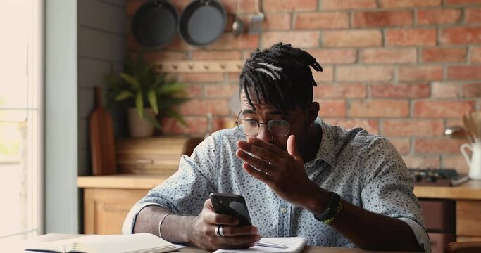 Awful news, negative emotions, bad message, personal life or business troubles concept. African hipster guy sits in kitchen with smartphone reading received sms bank notice feels stressed looks upset