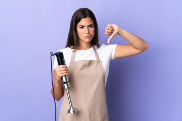 Brazilian woman using hand blender isolated on purple background showing thumb down with negative expression