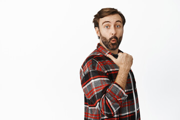 Wow check this out. Excited bearded man pulling surprised face, pointing at product behind his back, showing advertisement, special offer, white background