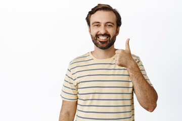 Smiling bearded man showing thumbs up in approval, like and agree, standing in t-shirt over white...