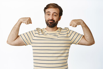 Image of sad funny guy showing his muscles, flexing biceps and grimacing upset with his body, standing over white background. Concept of workout and gym