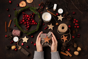 Yule winter solstice concept. Hands holding gift wrapped in linen fabric, tree branches, red berries, candles on wooden background. - obrazy, fototapety, plakaty