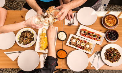 Set of tapas, salads and stews typical of Spanish food on a wooden table in which the hands of some people make a toast in a restaurant bar in Spain.