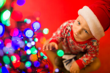 Fototapeta na wymiar baby in santa claus hat on a red background looks at the colorful lights from garlands.