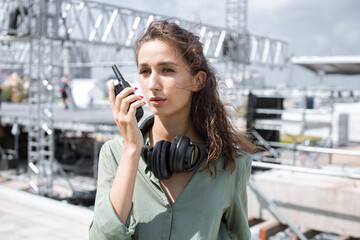 Installation of stage equipment and preparing for a live concert open air. Event manager portrait. Summer music city festival. Young serious woman speaks the walkie-talkie. - 470278365