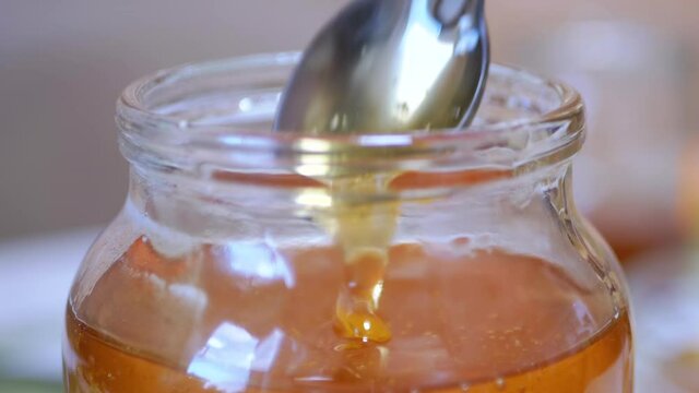 A Female Hand Picks Up with a Spoon Transparent, Liquid Honey from a Glass Jar. Yellow thick flow bee honey pouring, falling smoothly stream in tank. Healing, organic beekeeping product. Zoom.
