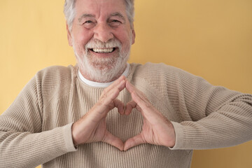 Portrait of happy senior bearded man making heart shape with his hands on yellow background
