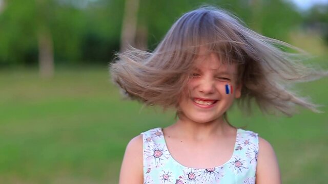 Child cheerful girl happily shakes head with her cheeks painted in the flag of France, her hair flying in different directions. Slow motion funny laughing little blonde kid girl 6-8 years old outdoor