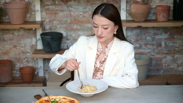 Portrait of stunning Chinese Asian woman in restaurant eating pasta and holding fork in her hand. Businesswoman in blouse enjoying lunch or dinner, old style industrial design cafe with clay pots