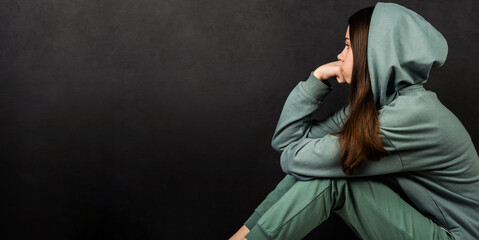 A teenage girl in a green sweatshirt sits alone and is sad against a dark background. Adolescent psychology concept. Emotions.