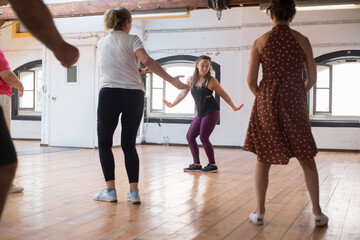 Happy young instructor teaching twist dance steps. Female Caucasian teacher with long fair hair demonstrating twist to her senior group in studio. Dance, hobby, healthy lifestyle concept