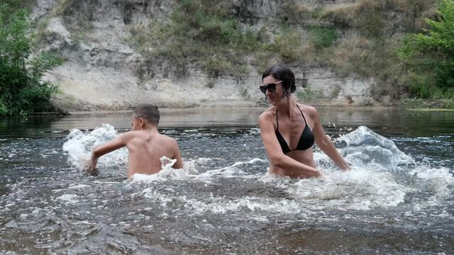 Happy Child and Mom are Splashing in a Shallow River, Creating Waves, Splashes. Water games in water with children on the beach. Concept of family, recreation, entertainment in nature. Slow motion.