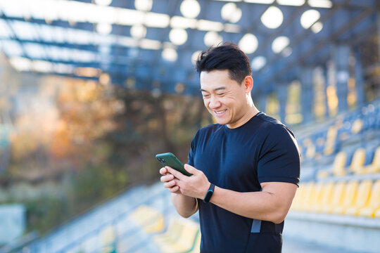 A healthy man watches a video on a mobile phone after a workout while sitting outdoors using an app on a 4g wireless device. Asian athlete is happy and smiling after running rest