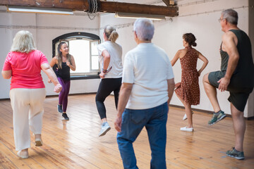 Hilarious young Caucasian instructor showing dance steps. Joyful female teacher with long hair in bright leggings demonstrating moves to her group of seniors. Dance, hobby, healthy lifestyle concept