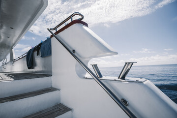 A fragment of a white safari diving boat with selective focus on chromium mooring bitts/bollards, wooden stairway leading to the deck of the vessel, and the railings; a warm sunny day, Maldives - obrazy, fototapety, plakaty