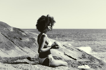 A side view of a ravishing young sensual African American woman with curly afro hair, sitting on the rock of the beach and drinking water from the coconut in her hands, black and white profile - obrazy, fototapety, plakaty