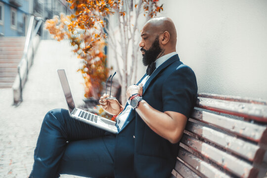 Side view of a serious mature bald elegant black businessman with a well-groomed beard, using his laptop while sitting on a wooden street bench with eyeglasses in his hand; African man using netbook - Powered by Adobe