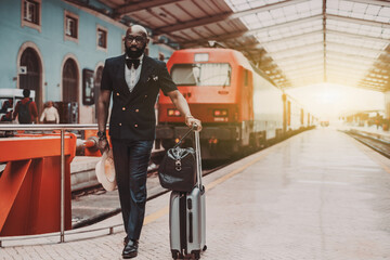 A dapper bearded bald black senior businessman in a fashionable suit with a vest and bowtie is standing on a platform of a railway station depot with his baggage, next to the red railroad train