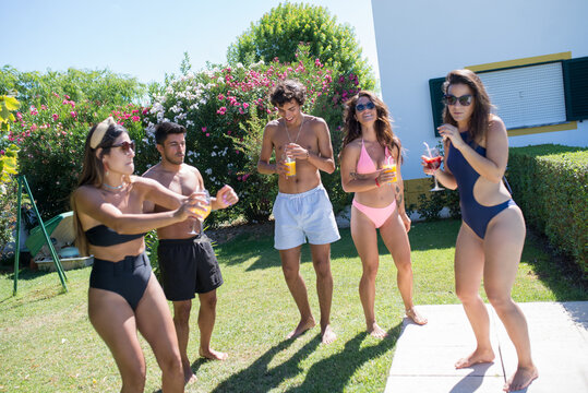 Merry friends partying at backyard. Young men and women in swimsuits standing near swimming pool, smiling, drinking cocktails, dancing. Leisure, friendship, party concept - Powered by Adobe