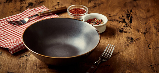 Empty dark bowl with fork, napkin and spices on wooden background