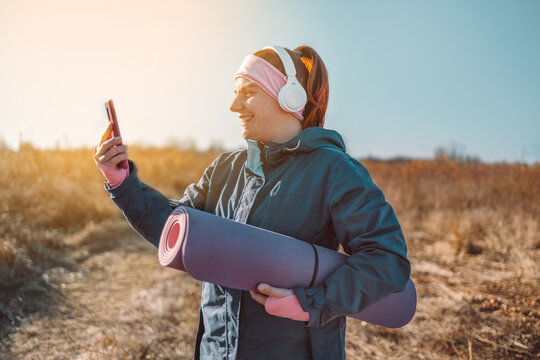 Young fitness woman holding yoga mat taking selfie photo after workout outdoors on a sunny day in nature