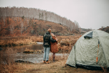 A loving couple resting in nature in the fall near the river and forest with a tent. Young woman in a beige hat, brown jacket, brown leggings. Young man in brown hat, khaki jacket, blue jeans