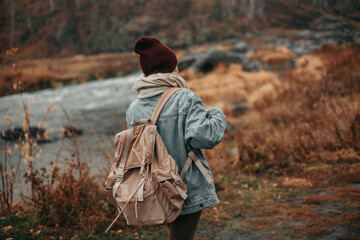 A young woman tourist blonde with long hair in a burgundy hat, warm blue jeans, brown leggings with a large beige backpack walks in the fall along the river. Back view.